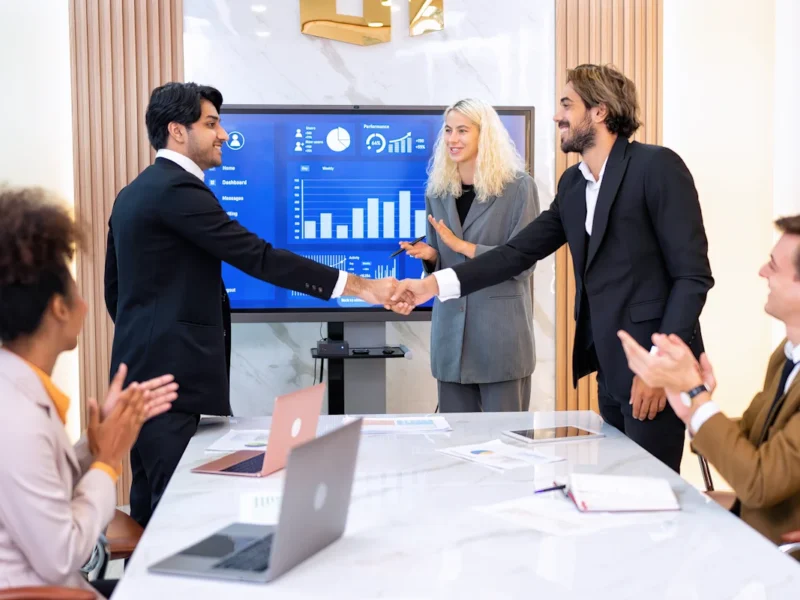 deux hommes qui se serrent la main devant un écran et un groupe de personne