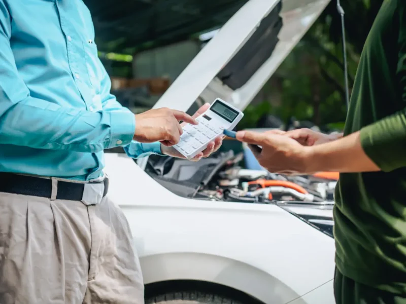 Un homme avec une machine à calculer qui discute avec un autre devant une voiture le capot ouvert